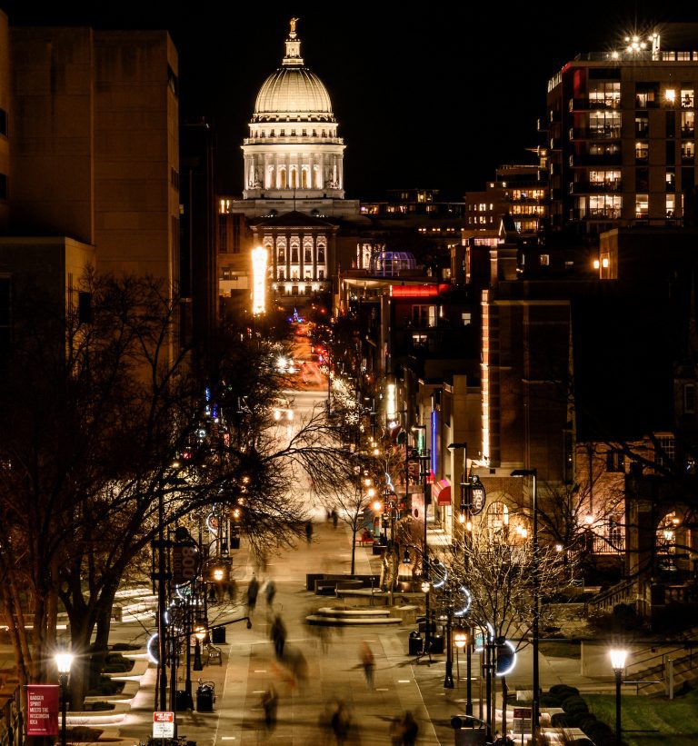 With the Wisconsin state capitol in the background, pedestrians and students walk along Library Mall and the State Street Mall at the University of Wisconsin-Madison after sunset